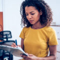 A Black businesswoman making notes on a clipboard inside of the office.