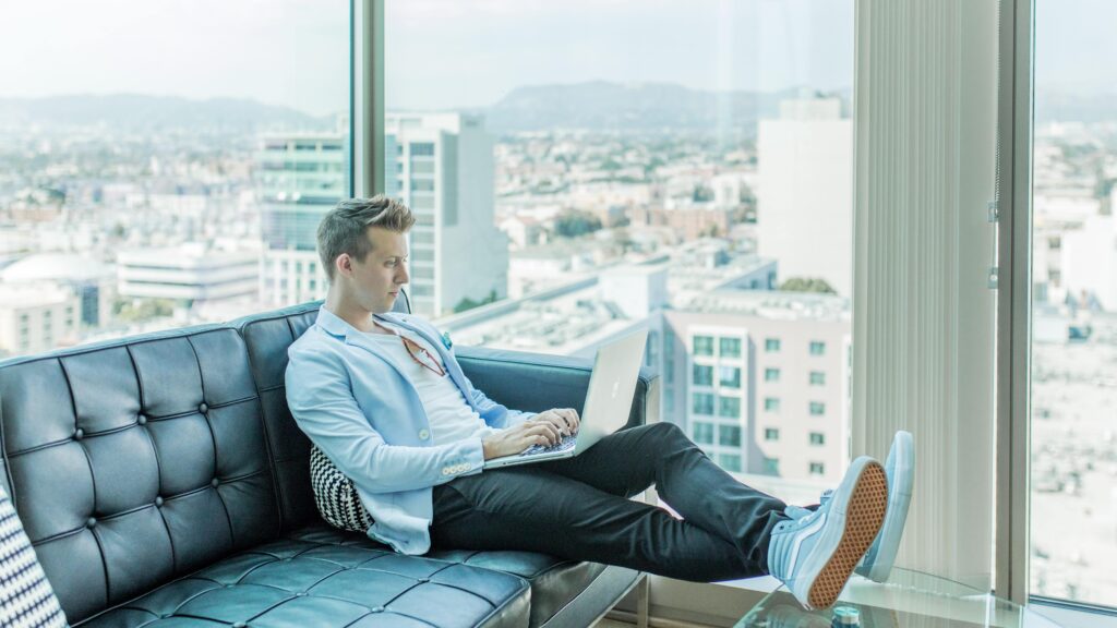 Photo of a young white man on a black couch working on a laptop with a window and city skyline in the background