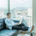 Photo of a young white man on a black couch working on a laptop with a window and city skyline in the background