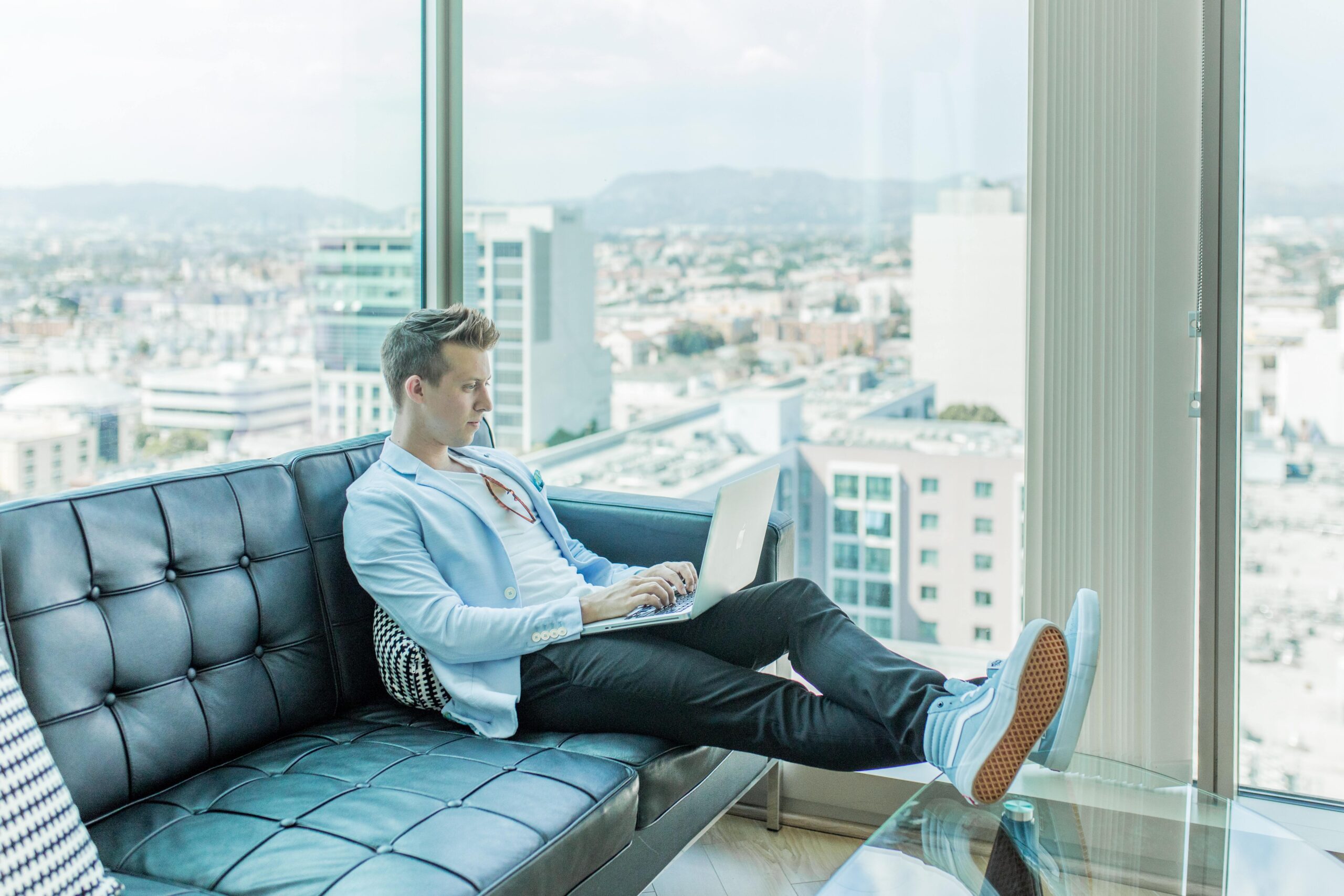 Photo of a young white man on a black couch working on a laptop with a window and city skyline in the background