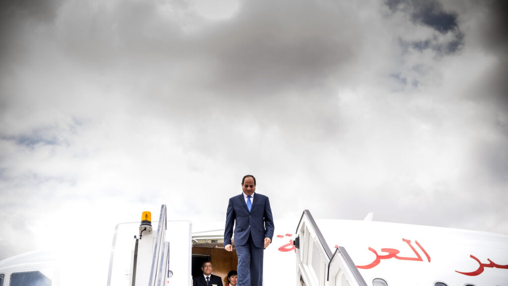 Egypt's President Abdel Fattah al-Sisi, wearing a blue suit and blue tie, exits a plane, standing at the top of a set of stairs that leads onto the tarmac.