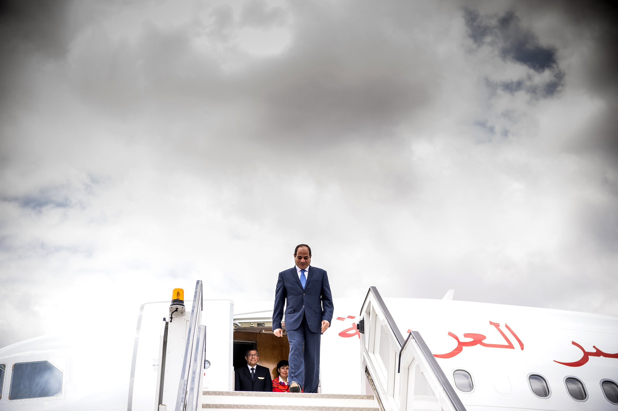 Egypt's President Abdel Fattah al-Sisi, wearing a blue suit and blue tie, exits a plane, standing at the top of a set of stairs that leads onto the tarmac.