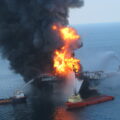 A ship in the ocean is obscured by with orange flames and black plumes of smoke. Three surrounding tugboats spray water in an attempt to put out the fire.