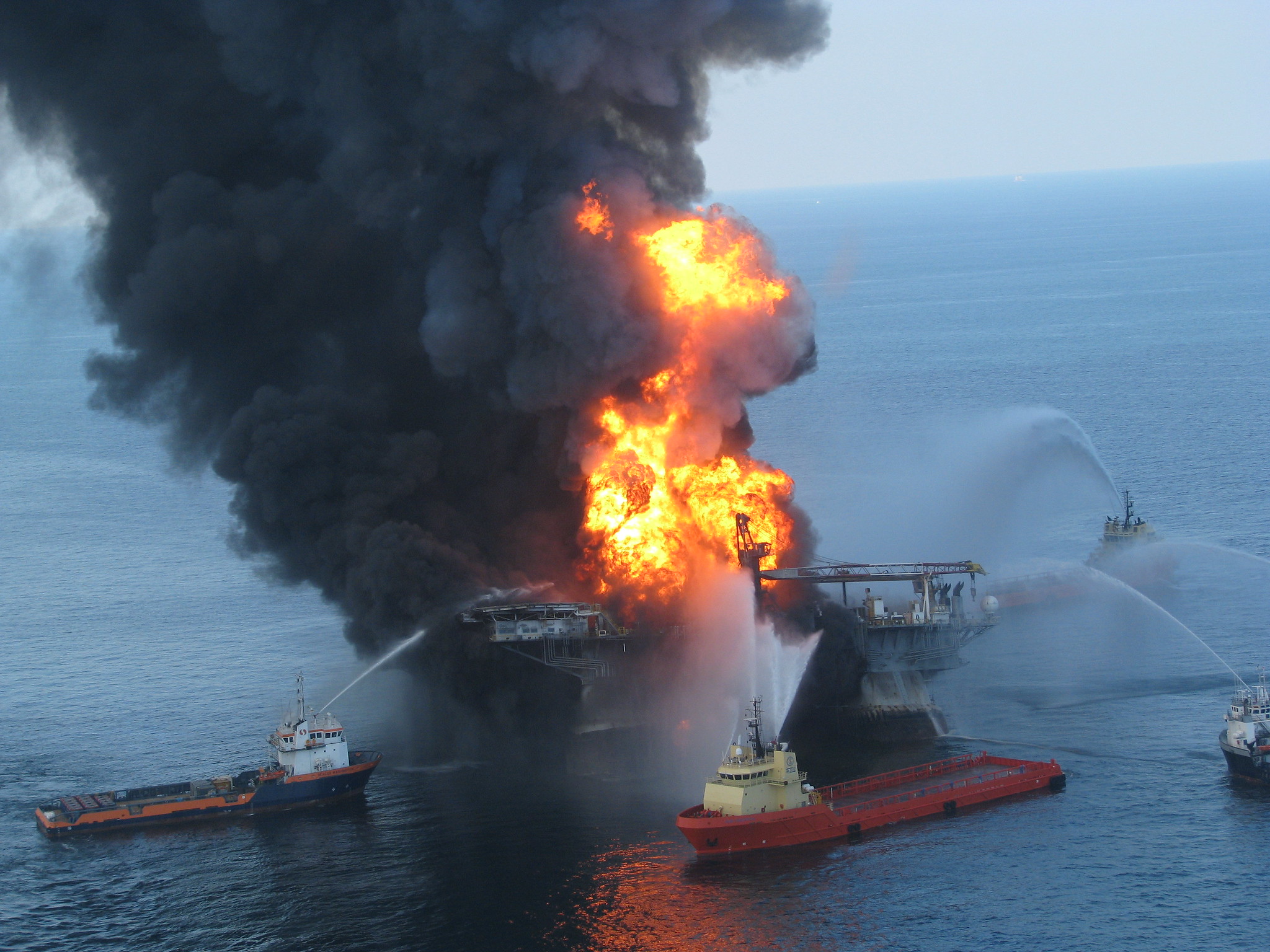 A ship in the ocean is obscured by with orange flames and black plumes of smoke. Three surrounding tugboats spray water in an attempt to put out the fire.