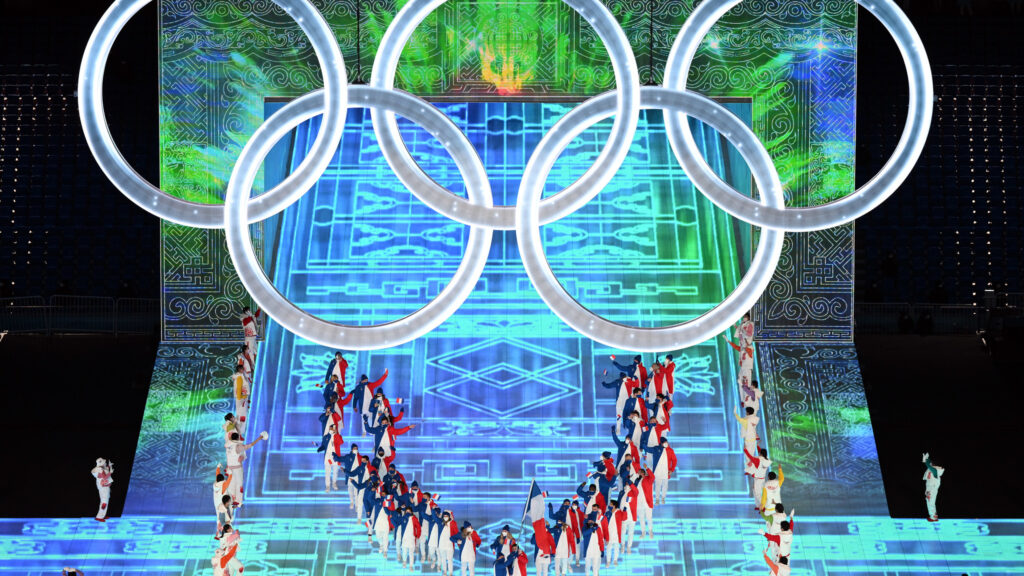 The opening ceremony for the winter Olympics in Beijing. The five Olympic rings hang over a stage lit in blue and green with athletes gathered in a semicircle underneath the rings