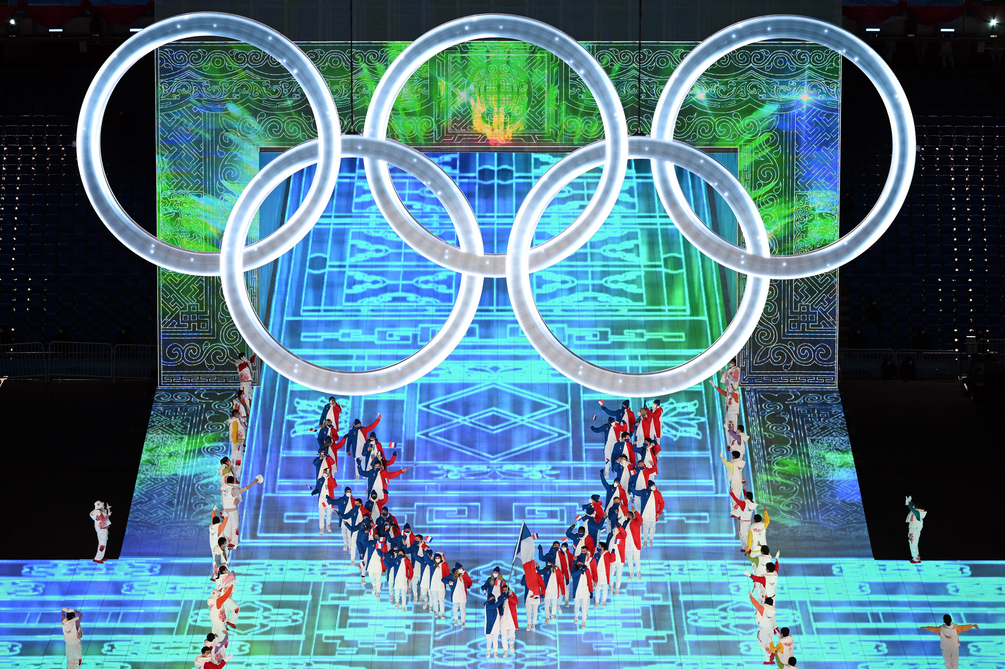 The opening ceremony for the winter Olympics in Beijing. The five Olympic rings hang over a stage lit in blue and green with athletes gathered in a semicircle underneath the rings