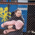 A woman holds her child, who wears a pink coat snowsuit. Both mother and child are behind a chain link fence.