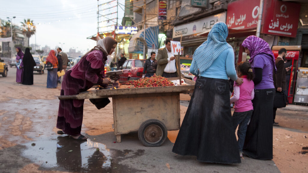 A woman sells strawberries from a cart on a rainy day. Two women and a little girl are clustered around the cart.