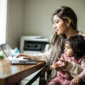 A woman sits with her young daughter in her lap, as she works on her laptop at a dining room table at home