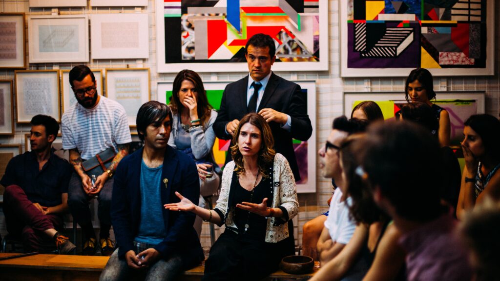 A group of people sit in two rows against a wall with colorful posters. They are looking at a central woman speaking.