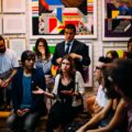 A group of people sit in two rows against a wall with colorful posters. They are looking at a central woman speaking.