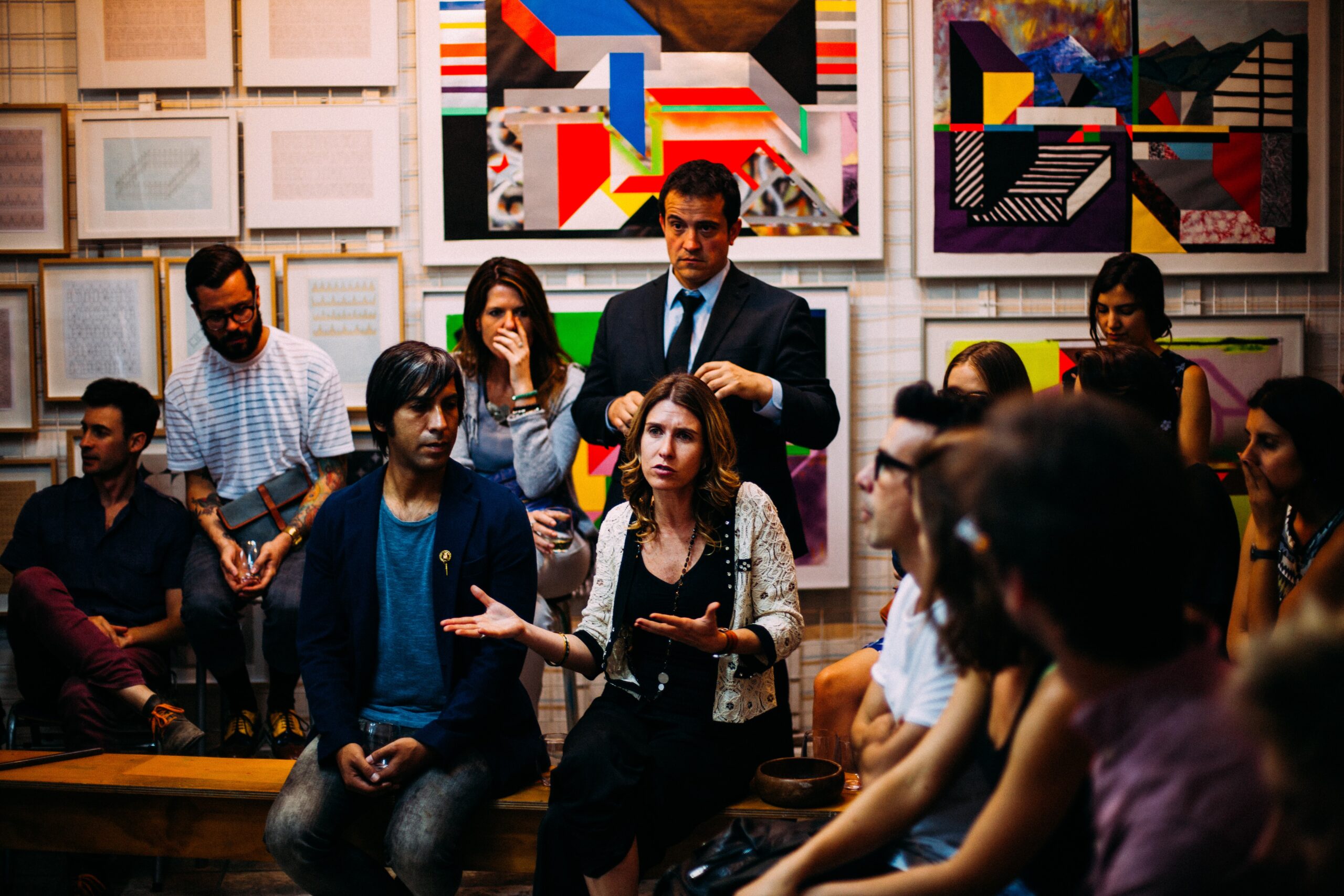A group of people sit in two rows against a wall with colorful posters. They are looking at a central woman speaking.