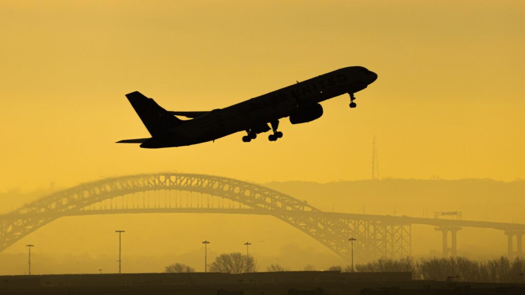 A silhouette of a plane taking off against a yellow sky with a bridge in the background