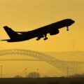 A silhouette of a plane taking off against a yellow sky with a bridge in the background