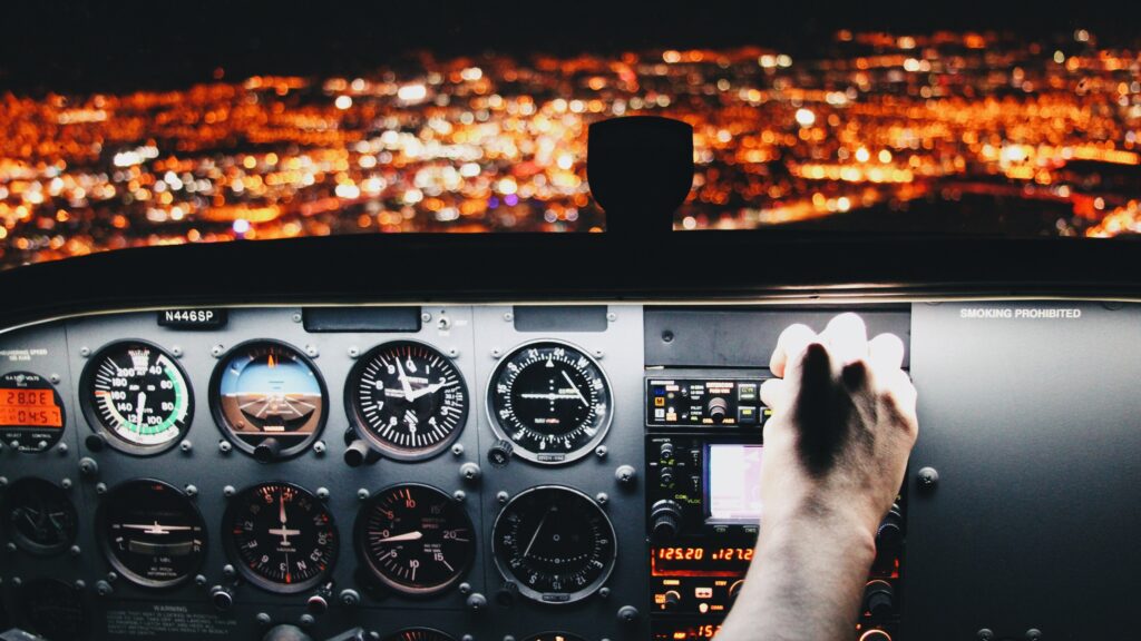 A view through a plane's front window at night, showing city lights. A man's hand adjusts the navigation controls.