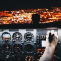 A view through a plane's front window at night, showing city lights. A man's hand adjusts the navigation controls.