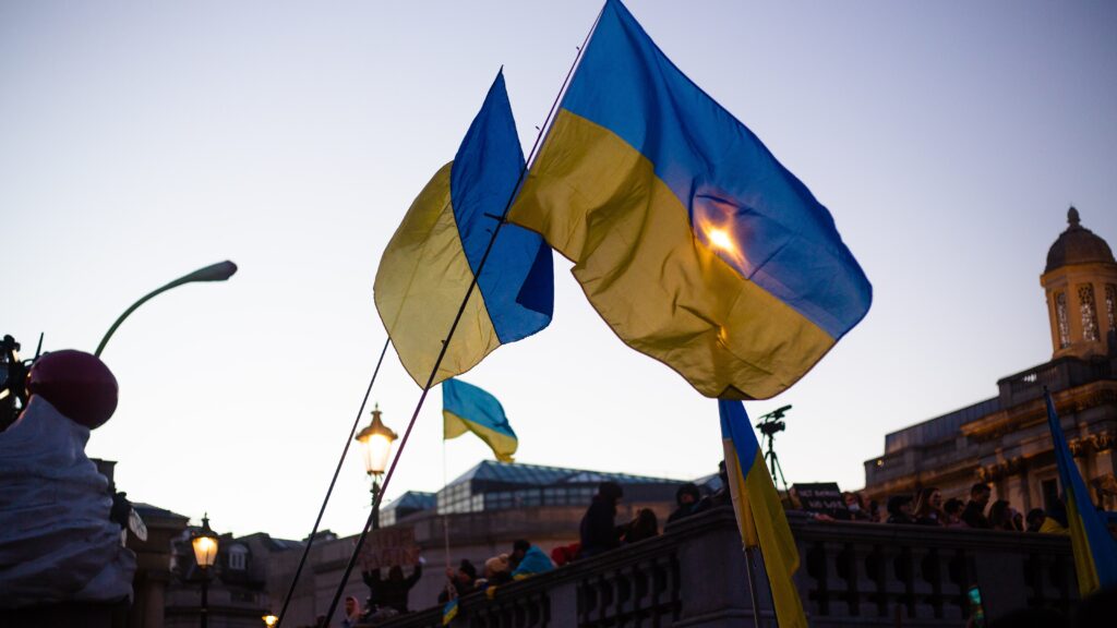 Two Ukrainian flags fly in the wind, with a crowd below them and buildings in the background