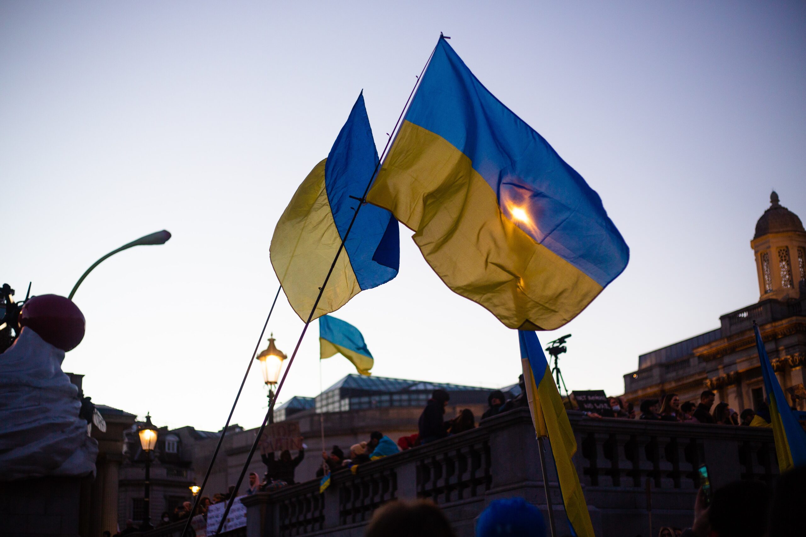 Two Ukrainian flags fly in the wind, with a crowd below them and buildings in the background