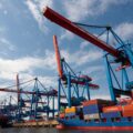 A cargo ship full of containers is docked in the water alongside a pier with cranes.