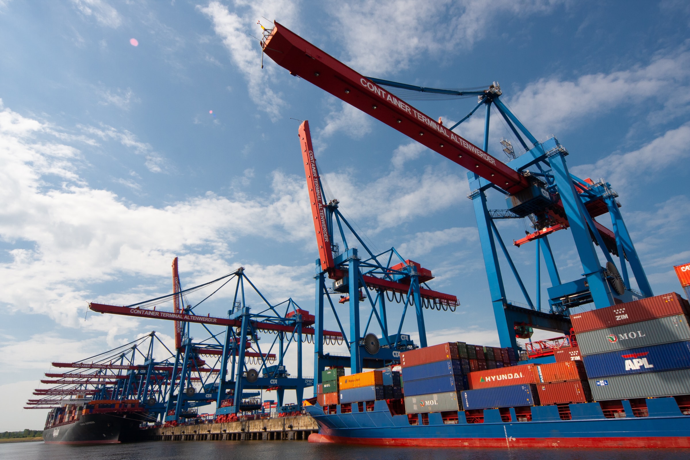 A cargo ship full of containers is docked in the water alongside a pier with cranes.