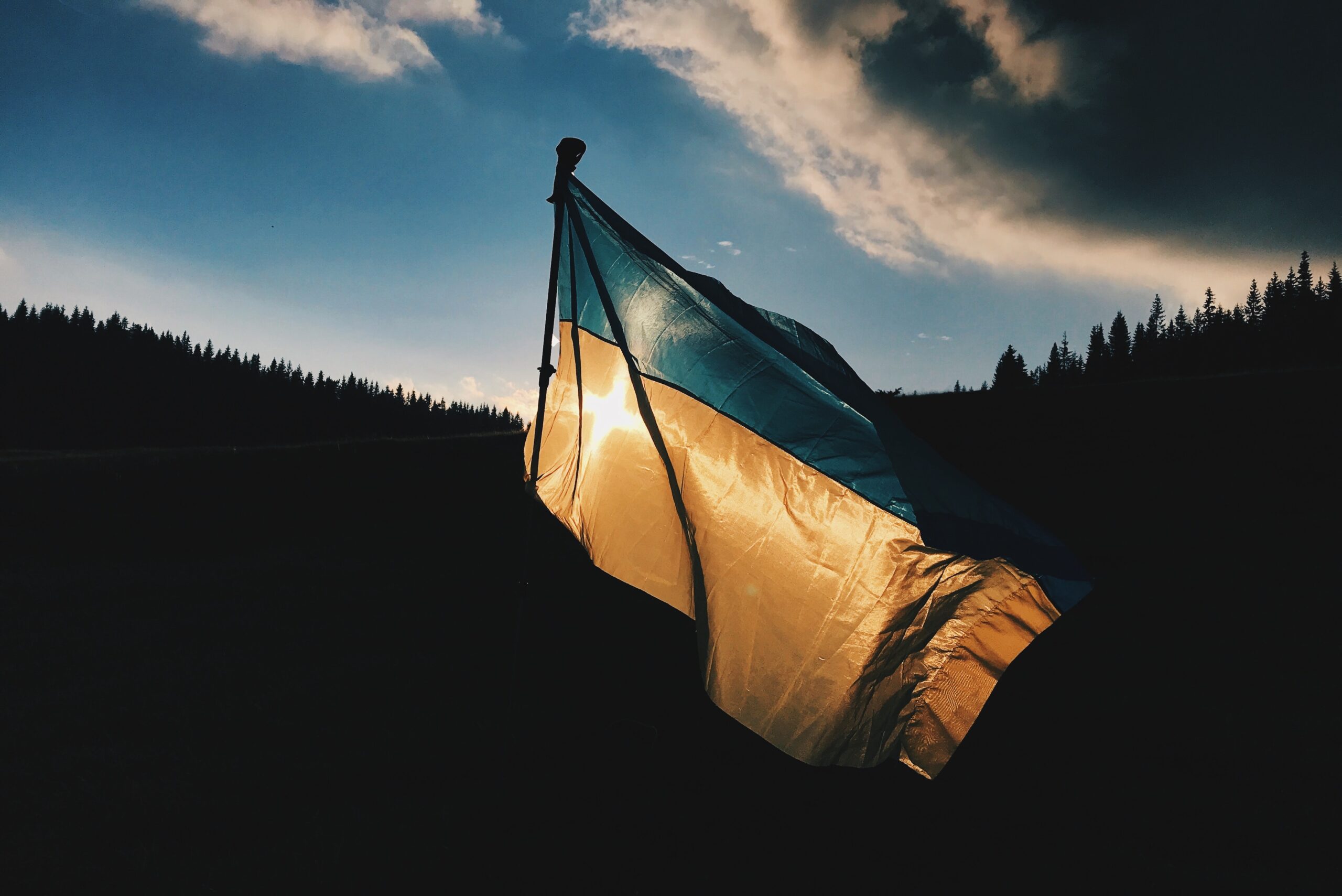 A Ukraine flag waves against a shadowed tree-filled hillside