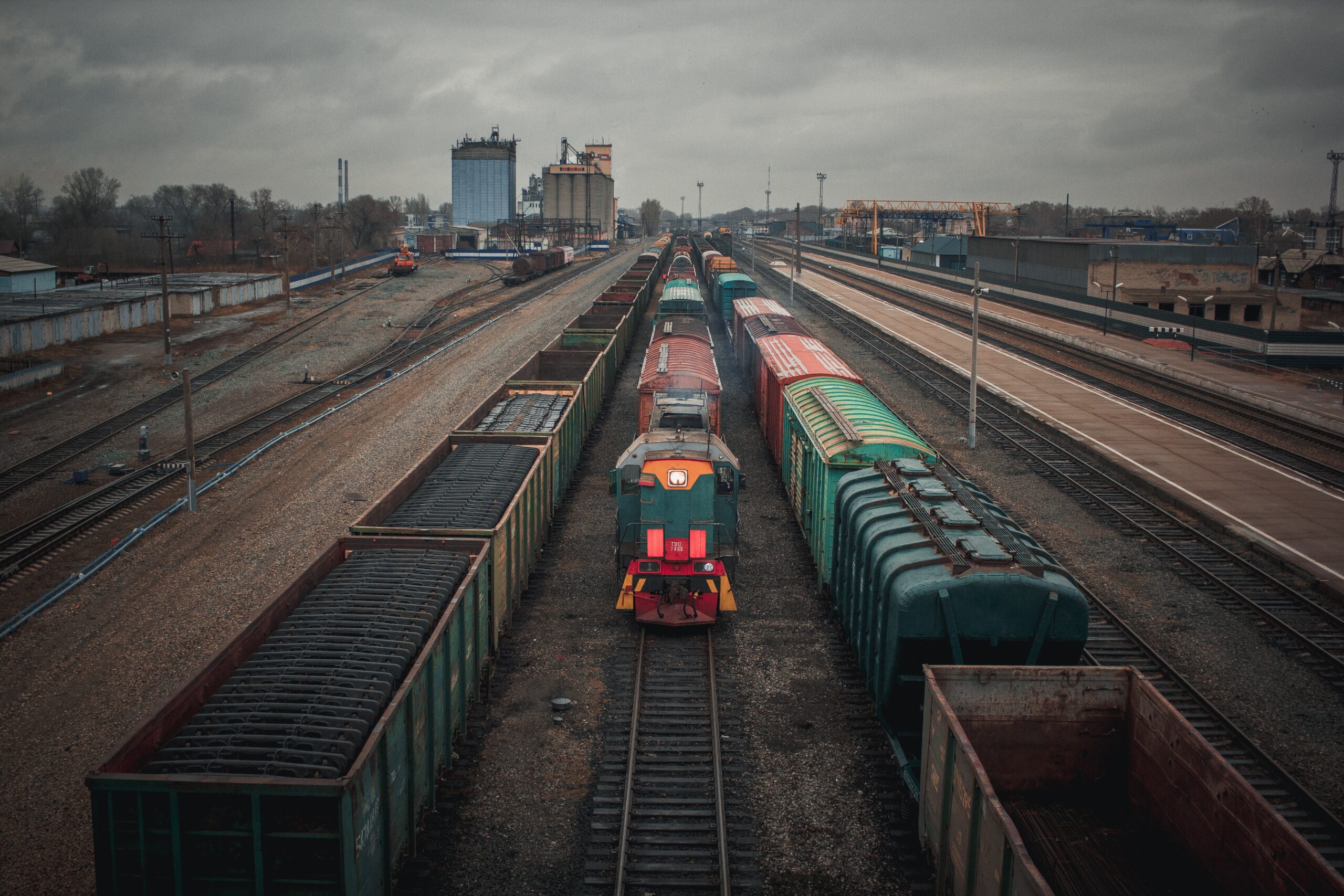 A train engine stands idle between two loaded freight trains in a train yard, on an overcast day