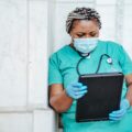 Image of a woman in teal scrubs leaning against a wall and looking at a tablet