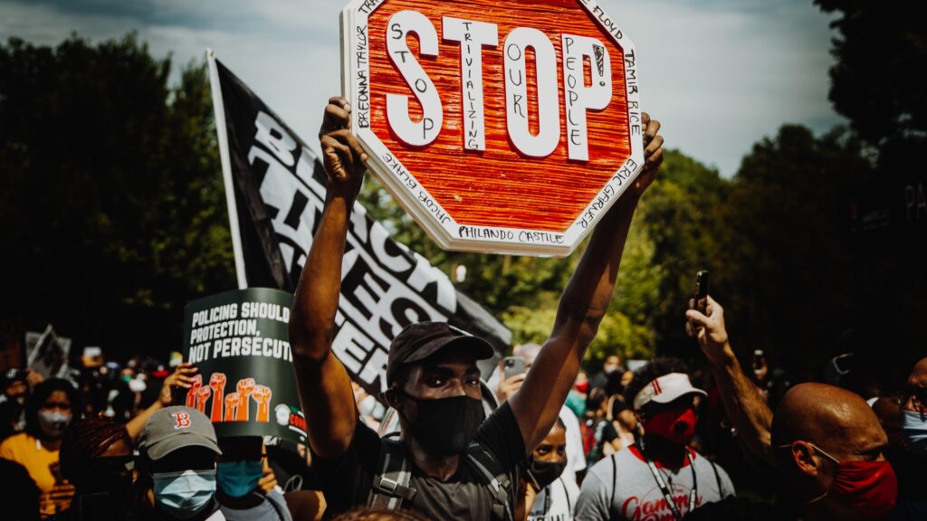 A man wearing a mask holds a stop sign with the names of Black Americans who have been killed by police. He is in the center of a crowd at a protest. Behind him is the Black Lives Matter flag.