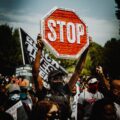 A man wearing a mask holds a stop sign with the names of Black Americans who have been killed by police. He is in the center of a crowd at a protest. Behind him is the Black Lives Matter flag.