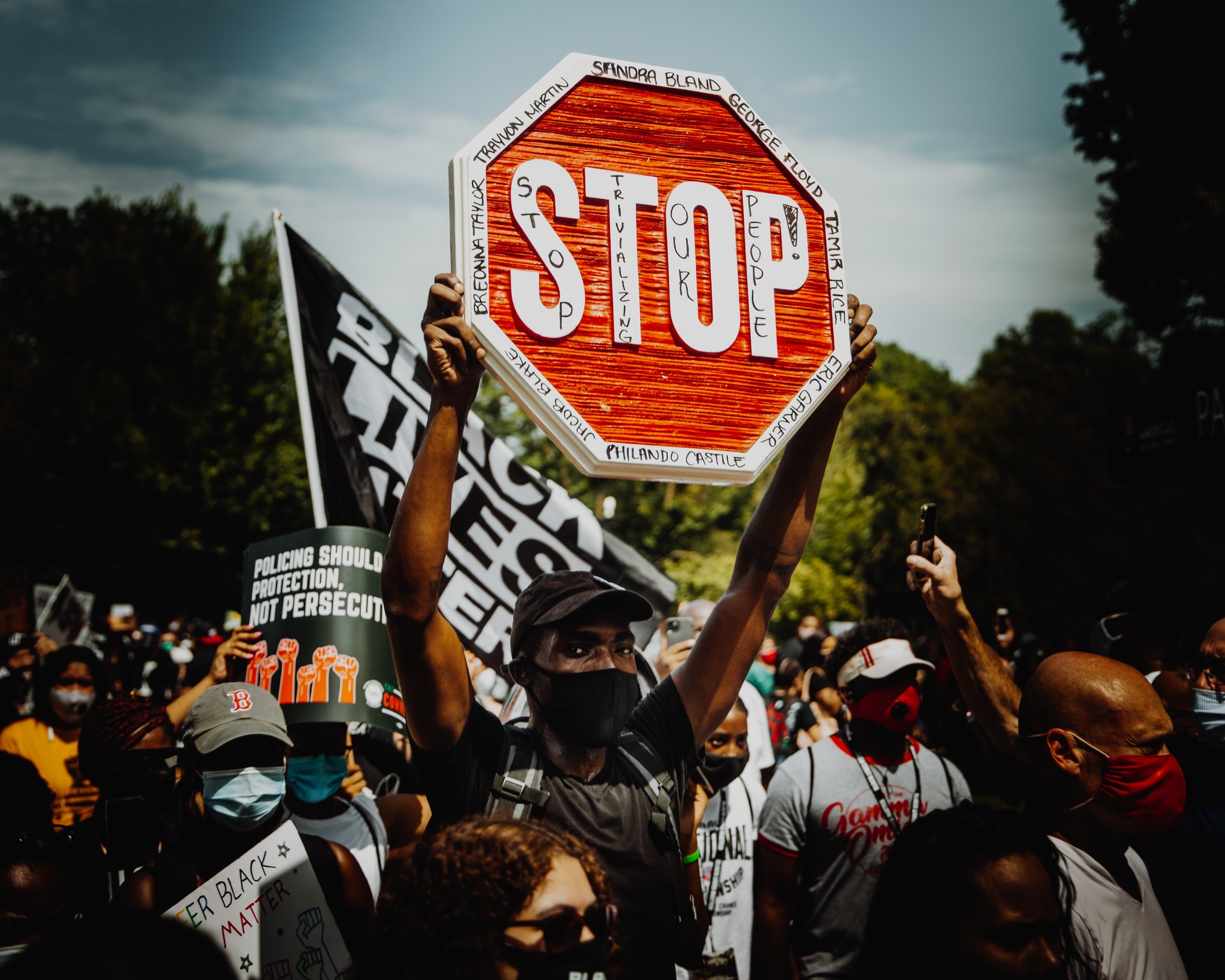 A man wearing a mask holds a stop sign with the names of Black Americans who have been killed by police. He is in the center of a crowd at a protest. Behind him is the Black Lives Matter flag.