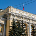 A view of a grand yellow building with a Russian flag and pine trees in front.