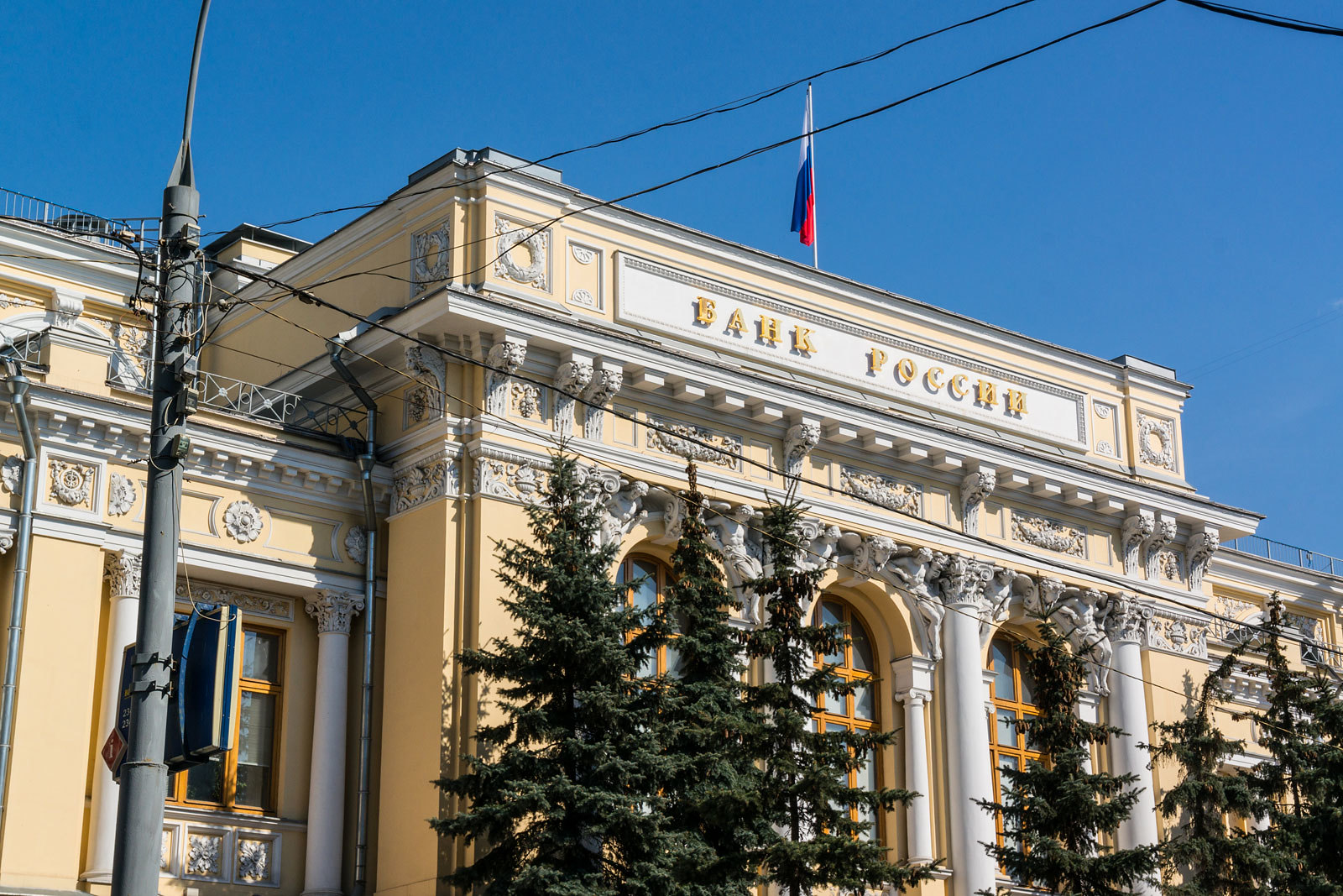 A view of a grand yellow building with a Russian flag and pine trees in front.