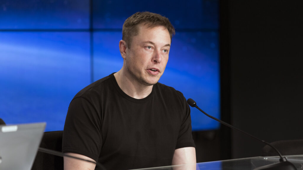 Elon Musk, wearing a black tshirt, sits at a table and speaks into a microphone. The background is blue.