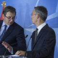 A man wearing a suit, blue tie, and glasses leans to talk to another grey-haired man in a suit standing behind a podium. the background is blue.