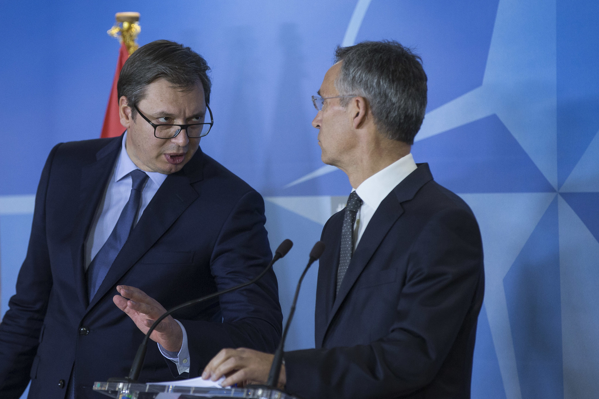 A man wearing a suit, blue tie, and glasses leans to talk to another grey-haired man in a suit standing behind a podium. the background is blue.
