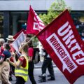 A rally in the street. A woman in a yellow safety vest carrying a large red flag that reads 