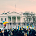 A crowd waving Ukrainian flags and holding signs protest the war gathers outside the fence of the White House, which can be seen in the background.