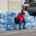 A woman sits on the sidewalk with her head buried in her hands. Around her are dozens of packs of bottle water.