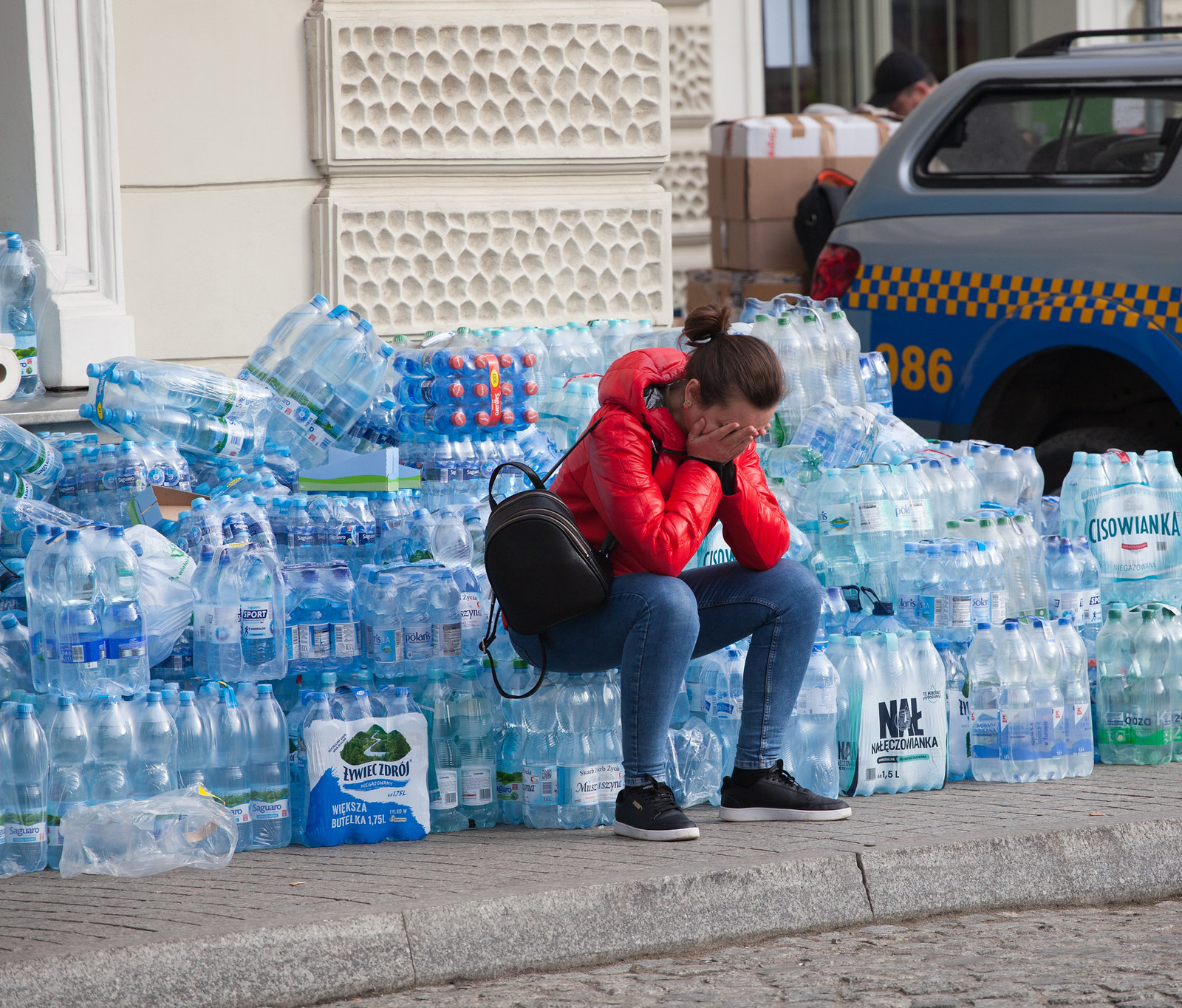 A woman sits on the sidewalk with her head buried in her hands. Around her are dozens of packs of bottle water.