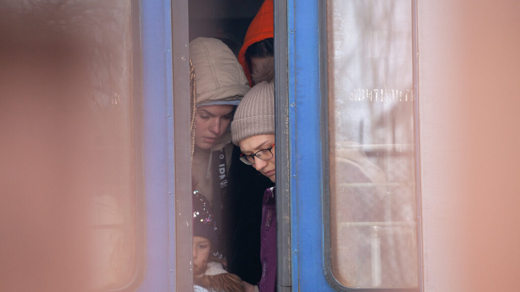Two women and a little girl in winter hats and coats are revealed by the slightly open doors of a train. A third person stands behind them in the train car, their face obscured by an orange hood.