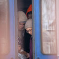 Two women and a little girl in winter hats and coats are revealed by the slightly open doors of a train. A third person stands behind them in the train car, their face obscured by an orange hood.