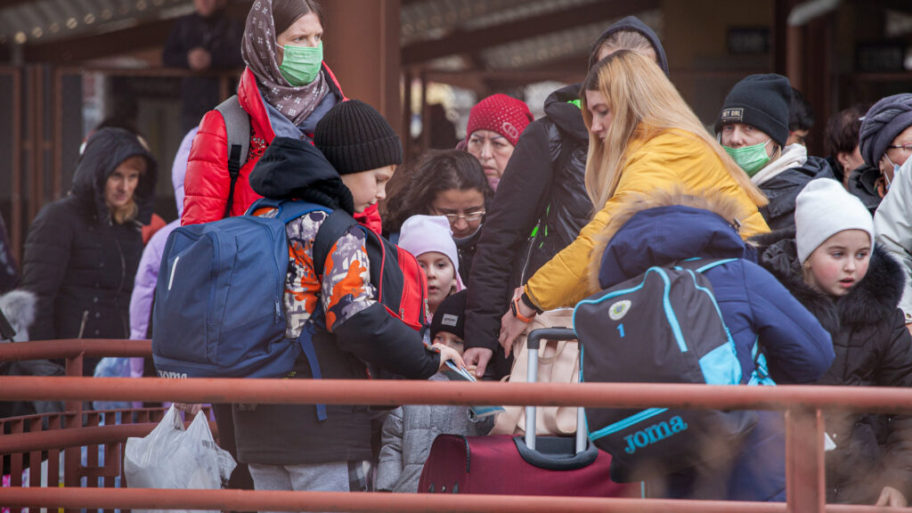 A crowd of women and children wearing winter clothing and masks, and carrying bags group together outside as they wait for a train.