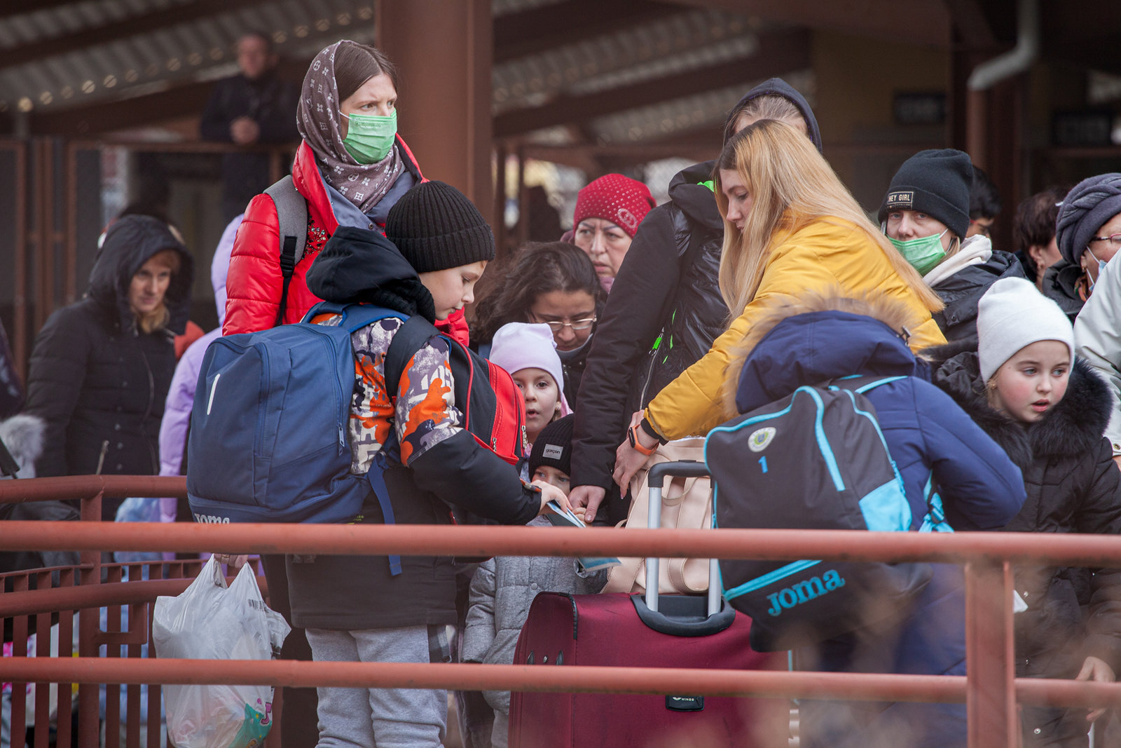 A crowd of women and children wearing winter clothing and masks, and carrying bags group together outside as they wait for a train.