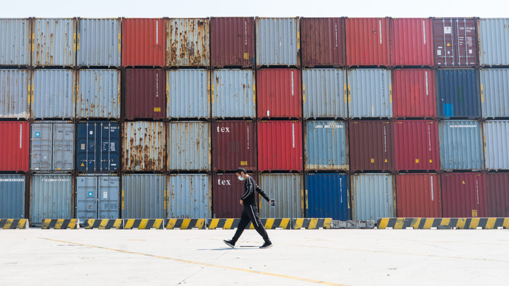A wide shot shows a man wearing black with a face mask walking past a tall stack of containers in Yangshan port in Shanghai, China.