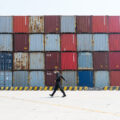 A wide shot shows a man wearing black with a face mask walking past a tall stack of containers in Yangshan port in Shanghai, China.