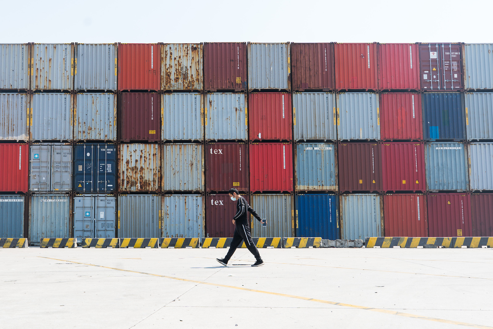 A wide shot shows a man wearing black with a face mask walking past a tall stack of containers in Yangshan port in Shanghai, China.