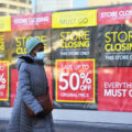 A woman wearing a grey winter coat, blue hat, and blue surgical face mask walks past a wall of black and yellow 