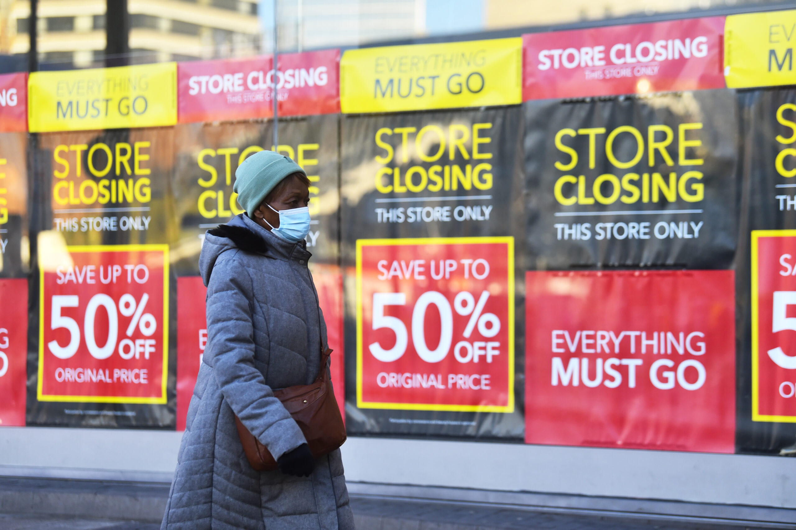 A woman wearing a grey winter coat, blue hat, and blue surgical face mask walks past a wall of black and yellow