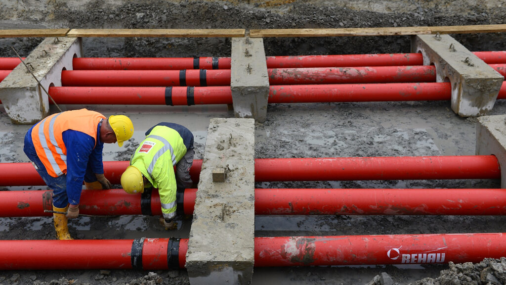 Two workers in yellow helmets and orange and yellow safety vests bend over six red pipes at a construction site.