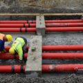 Two workers in yellow helmets and orange and yellow safety vests bend over six red pipes at a construction site.